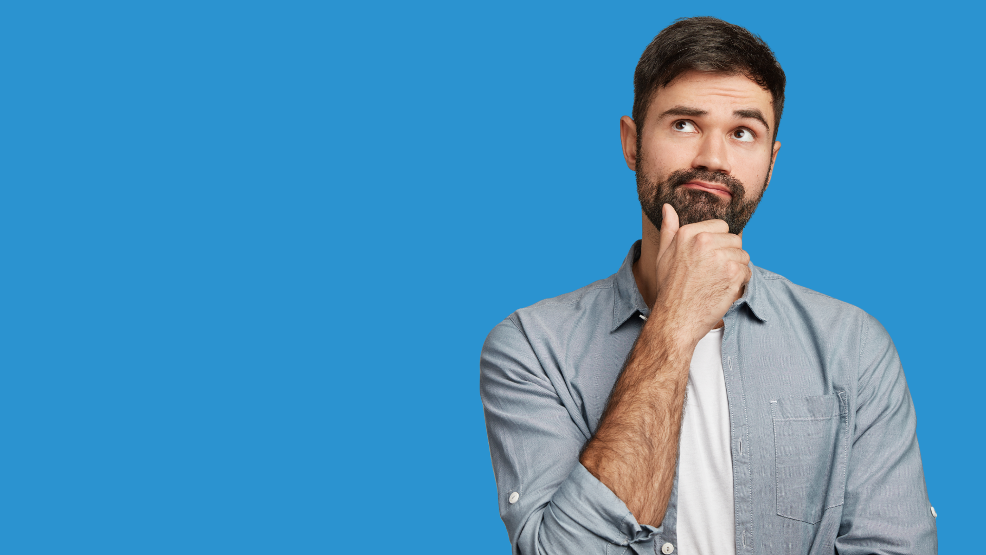 Homem jovem de barba, com expressão pensativa e a mão no queixo, olhando para cima. Ele veste camisa azul clara e está em frente a um fundo azul sólido.