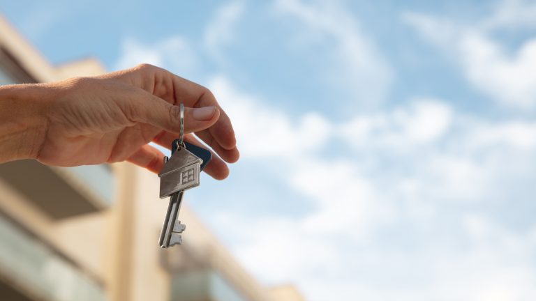 Mão segurando uma chave com pingente de casa, simbolizando a conquista ou garantia de um imóvel, com fundo desfocado de um prédio e céu azul.