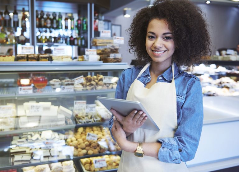 Mulher jovem usando avental e segurando um tablet enquanto sorri em uma padaria, com vitrines cheias de doces e produtos ao fundo.