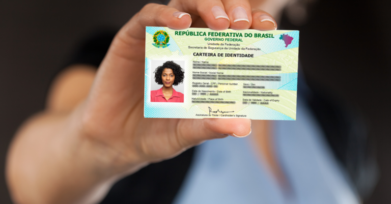 Pessoa segurando uma carteira de identidade brasileira (RG) em destaque, com a mão estendida para a câmera. O documento está em foco, mostrando foto e dados pessoais, enquanto o fundo permanece desfocado.