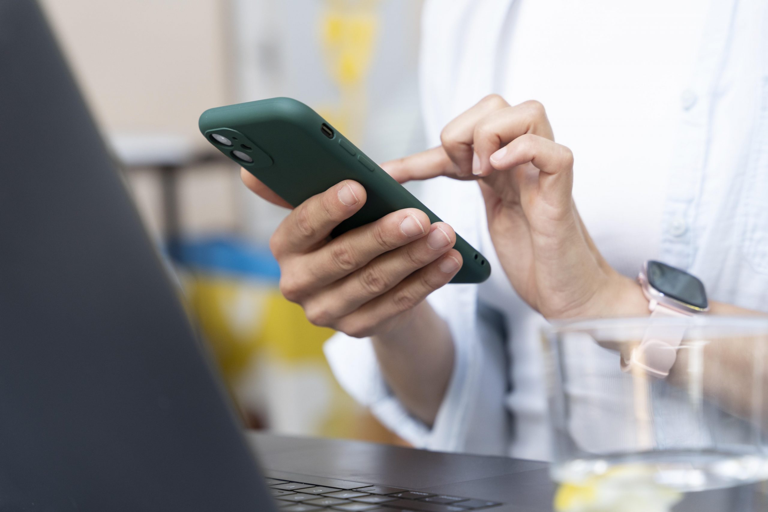 Pessoa usando um smartphone enquanto trabalha em frente a um notebook, representando o uso de aplicativos bancários ou transações online.