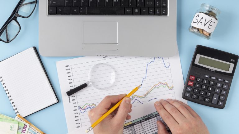 Pessoa analisando gráficos financeiros sobre uma mesa, com laptop, calculadora, lupa e um pote marcado como "save", representando organização e planejamento financeiro.