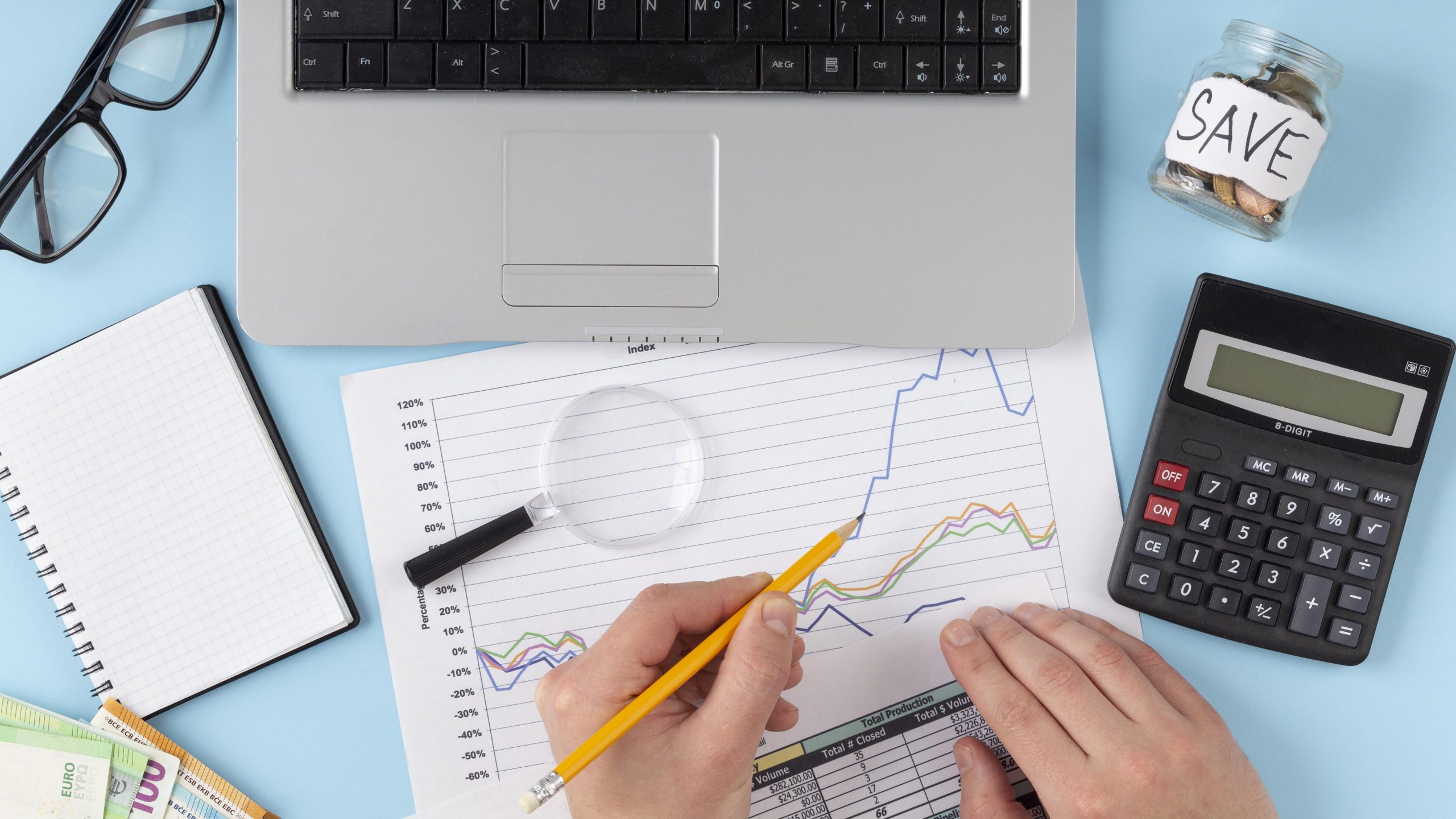 Pessoa analisando gráficos financeiros sobre uma mesa, com laptop, calculadora, lupa e um pote marcado como "save", representando organização e planejamento financeiro.
