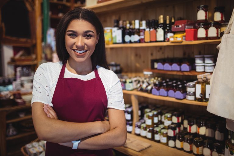 Mulher empreendedora sorrindo em seu pequeno comércio, usando avental, com prateleiras de produtos ao fundo.
