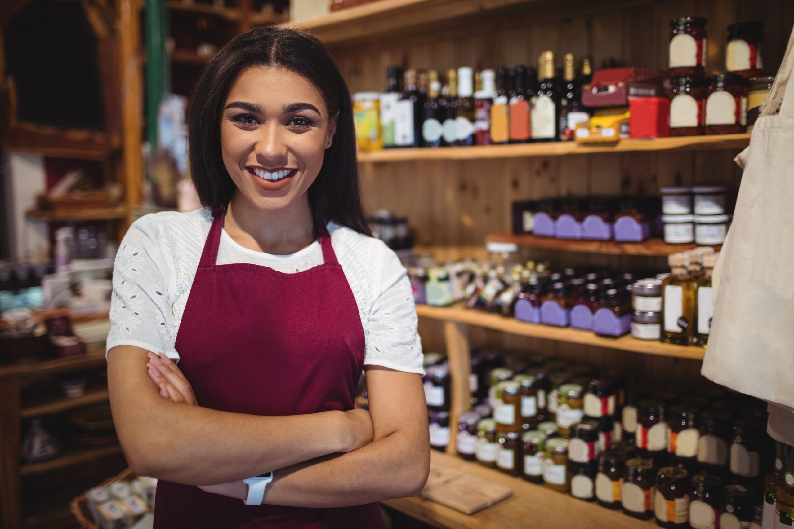 Mulher empreendedora sorrindo em seu pequeno comércio, usando avental, com prateleiras de produtos ao fundo.