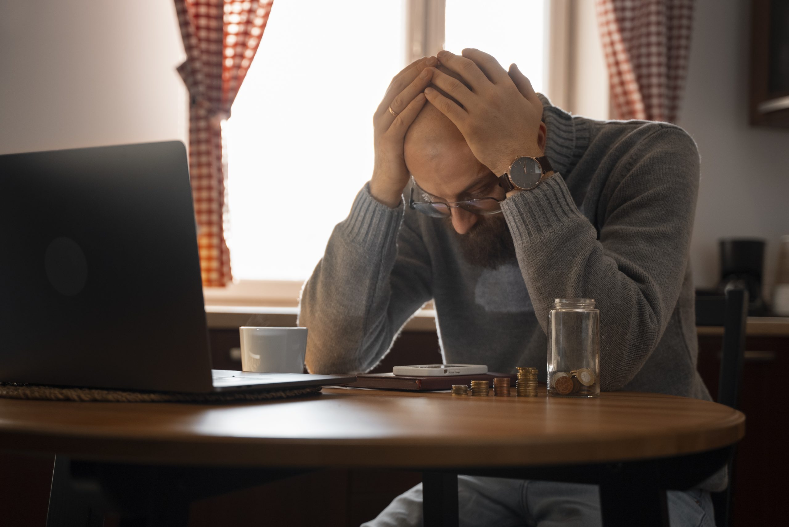Homem sentado à mesa com a cabeça apoiada nas mãos, diante de notebook e contas, demonstrando preocupação com dívidas financeiras.