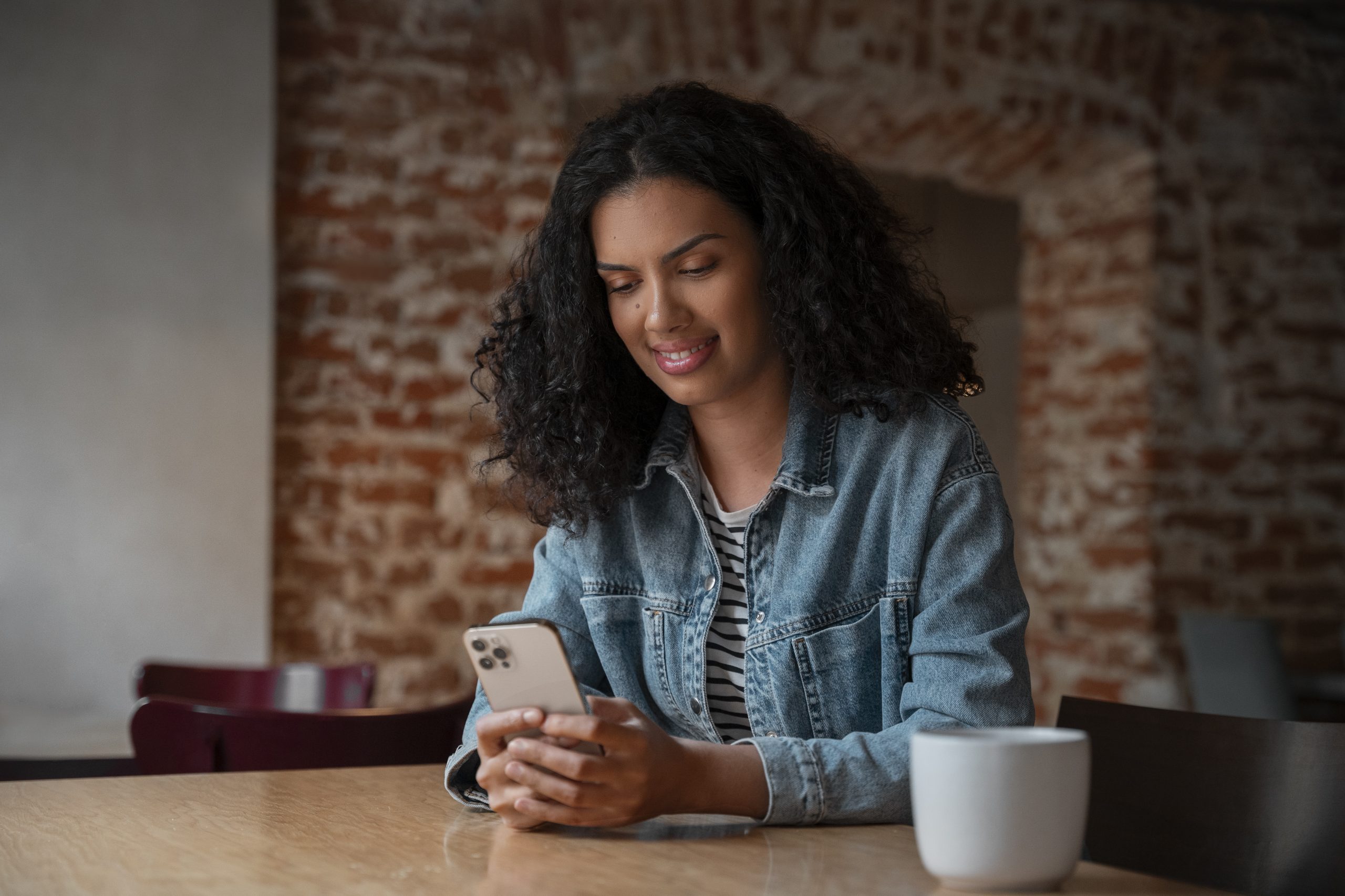 Mulher utilizando o celular sentada à mesa, com uma xícara de café ao lado, representando uso de banco digital e organização financeira.