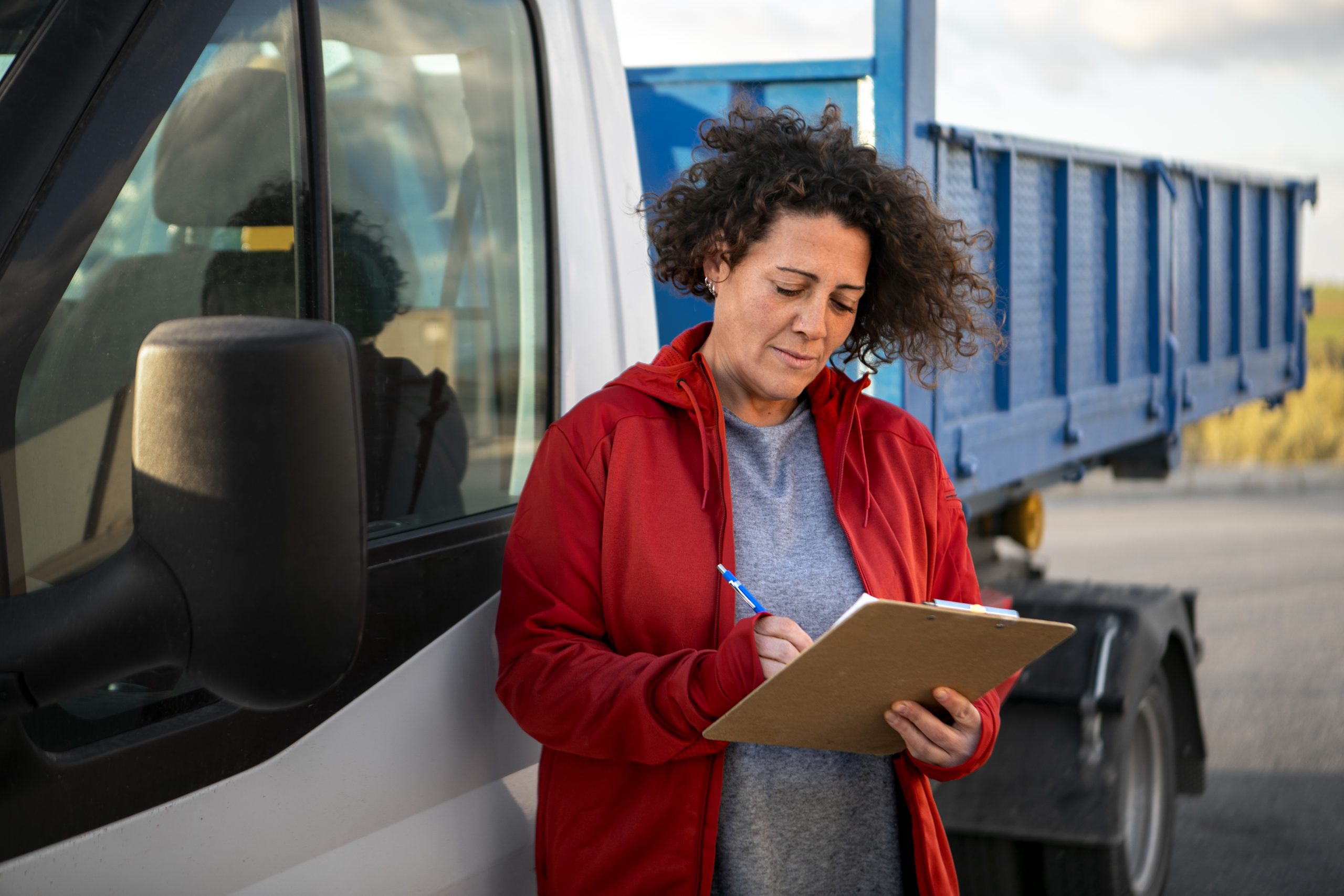 Mulher escrevendo em prancheta ao lado de caminhão, representando organização e controle financeiro no setor de transporte.