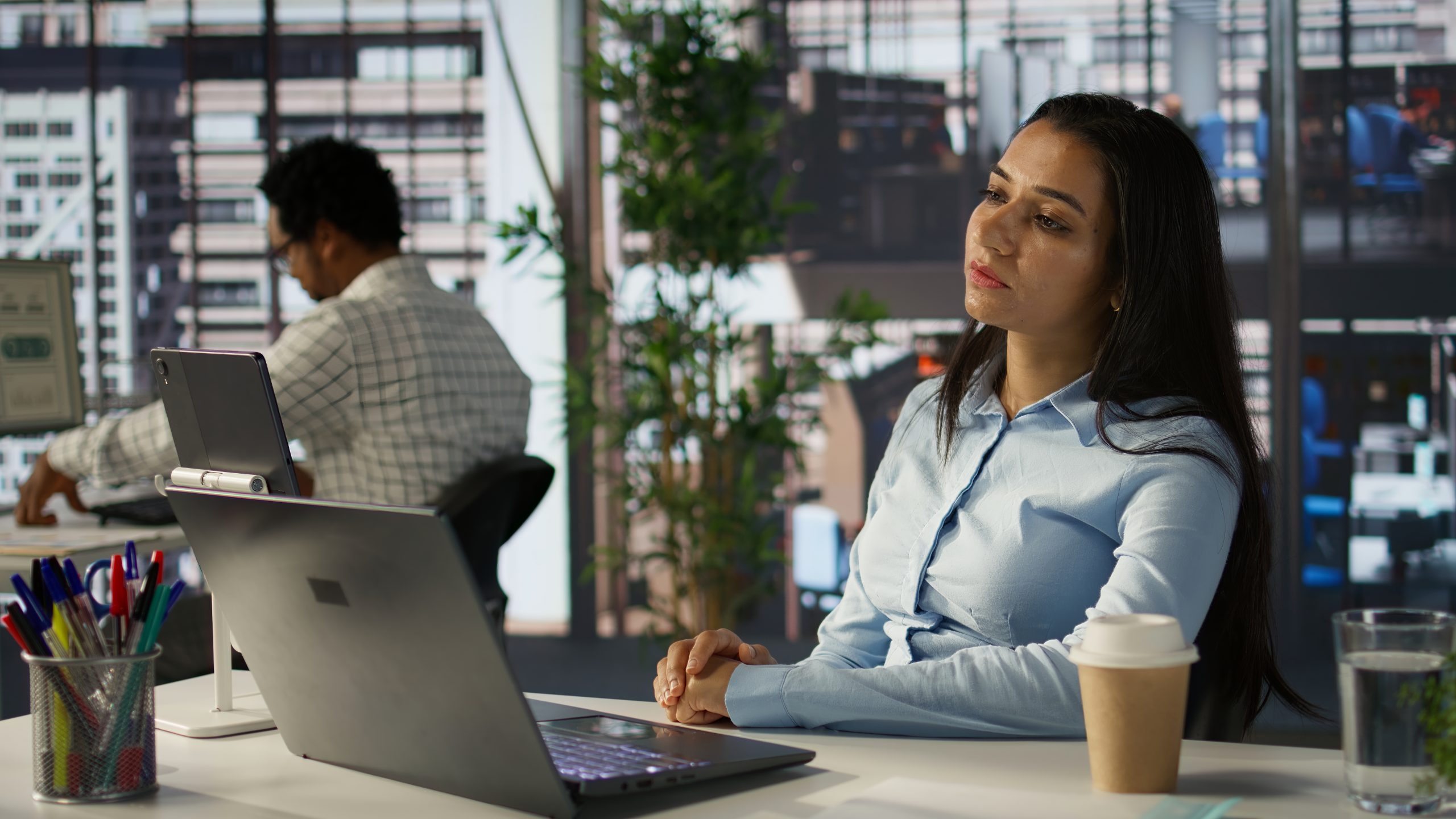 Mulher sentada em mesa de escritório usando notebook, com copo de café ao lado, enquanto colega trabalha ao fundo em ambiente corporativo com janelas amplas e vista urbana.