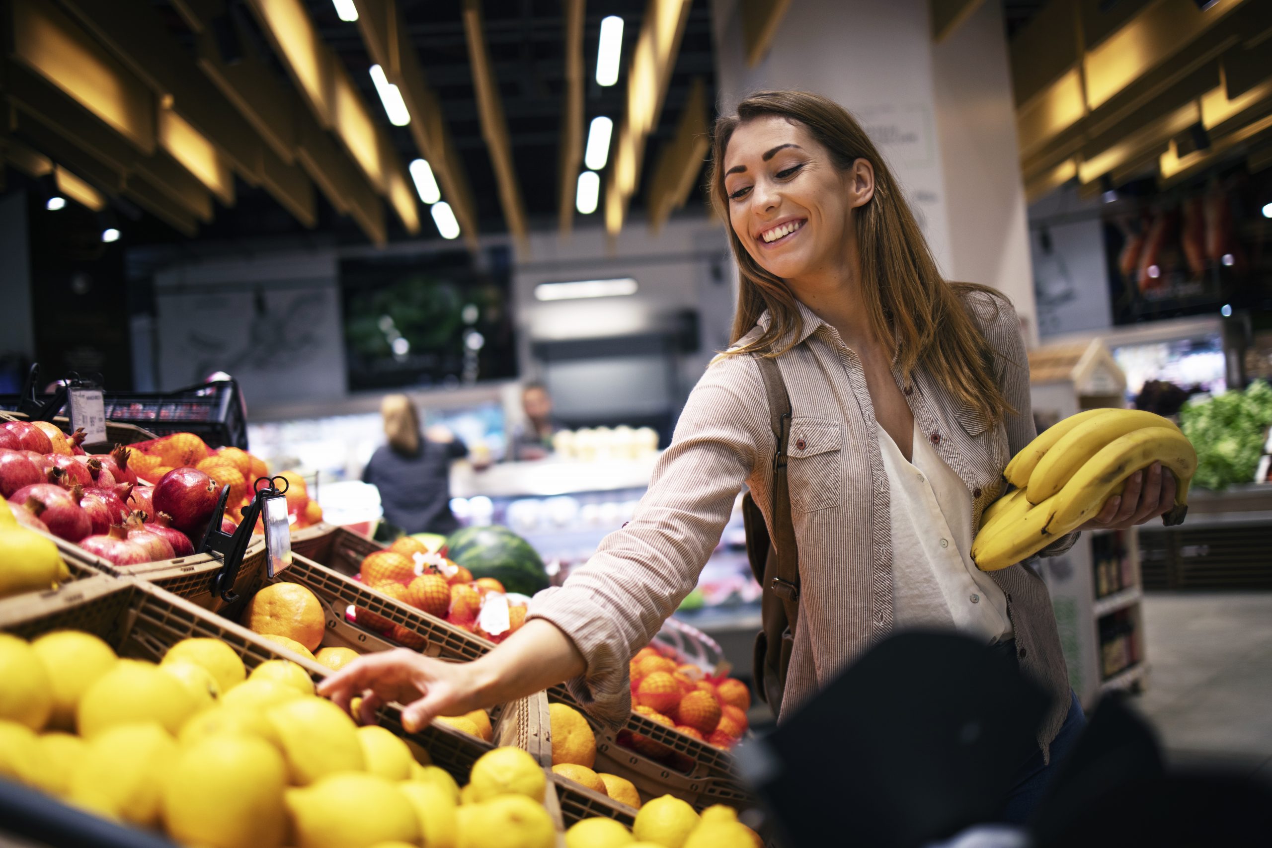 Mulher escolhendo frutas em um supermercado, segurando bananas enquanto pega maçãs na banca, em ambiente iluminado com prateleiras de alimentos ao fundo, representando compras no mercado e planejamento doméstico.