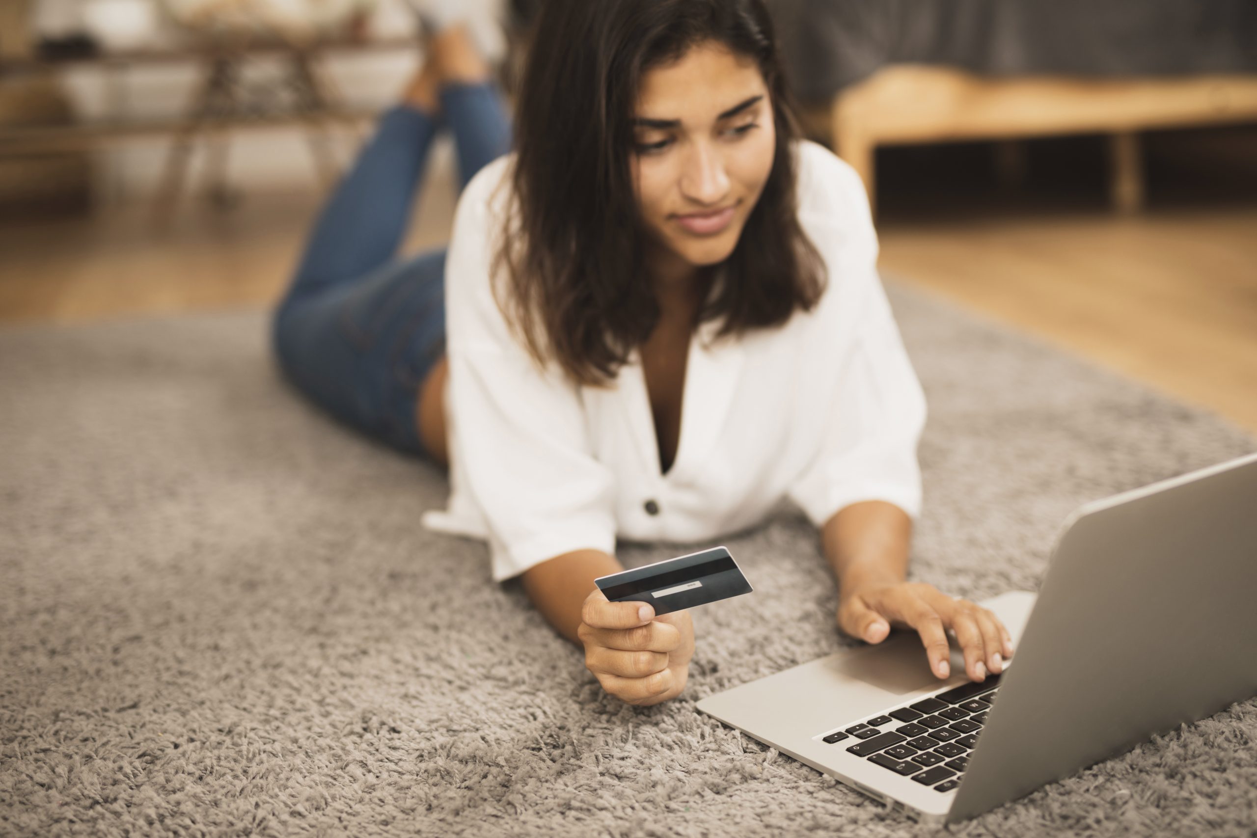 Mulher deitada no tapete usando notebook enquanto segura cartão de crédito, representando compras online, controle financeiro ou uso consciente do cartão.