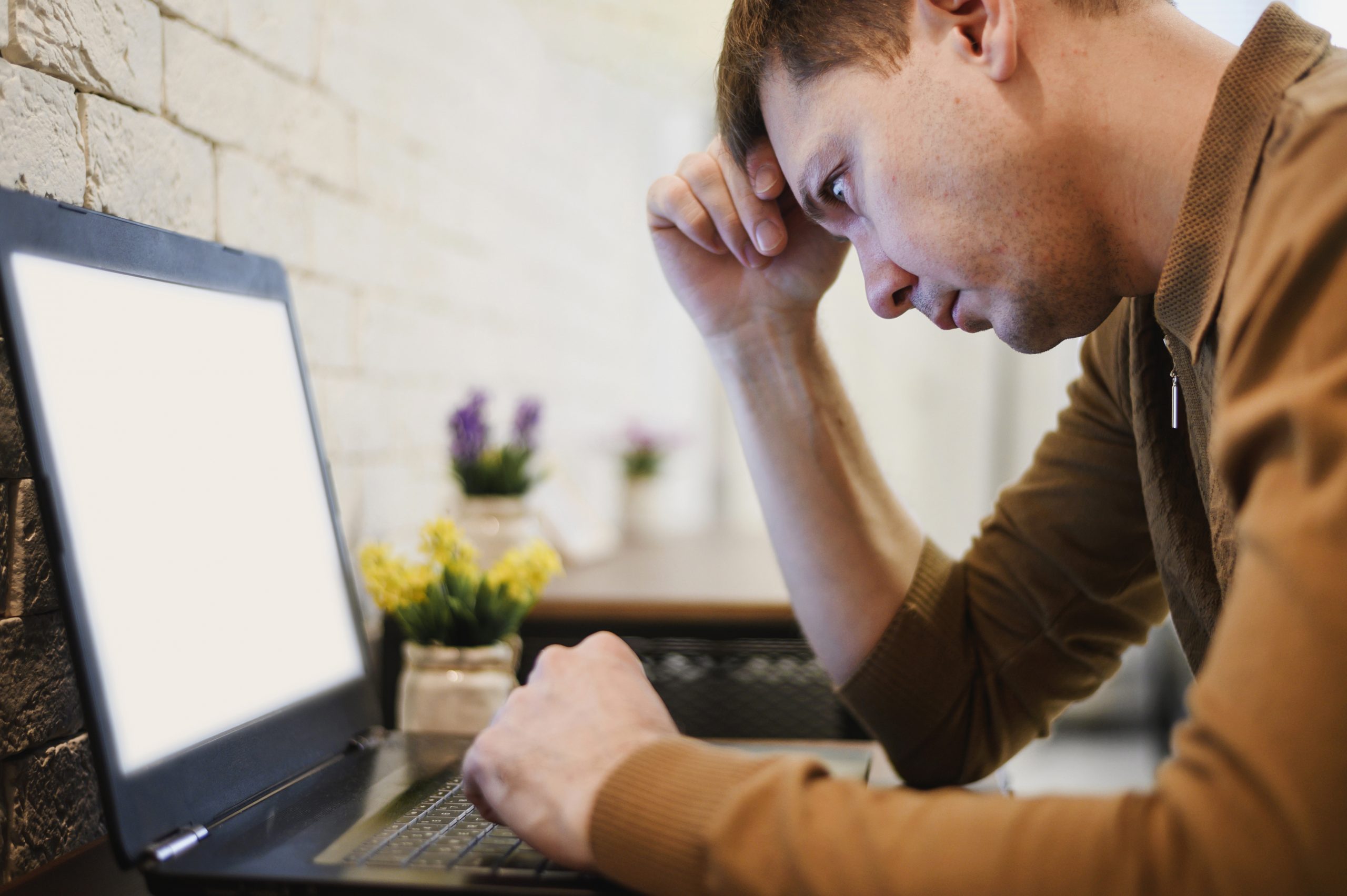 Homem jovem sentado à mesa, olhando fixamente para a tela do notebook com expressão preocupada, apoiando a mão na testa. Ao fundo, parede clara e pequenos vasos de plantas sobre a mesa.