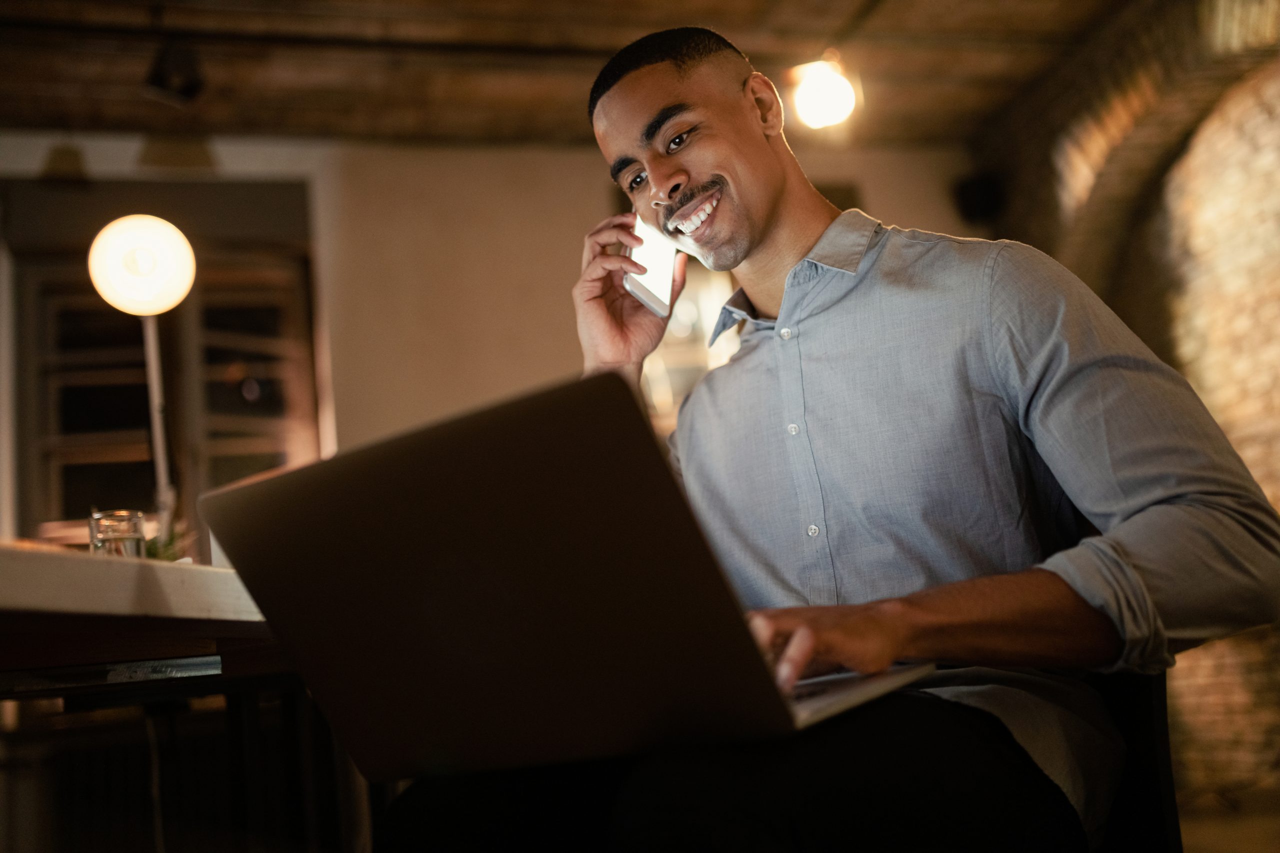 Homem sorrindo enquanto fala ao telefone e usa notebook em ambiente interno iluminado, representando organização financeira, trabalho remoto ou atendimento digital.